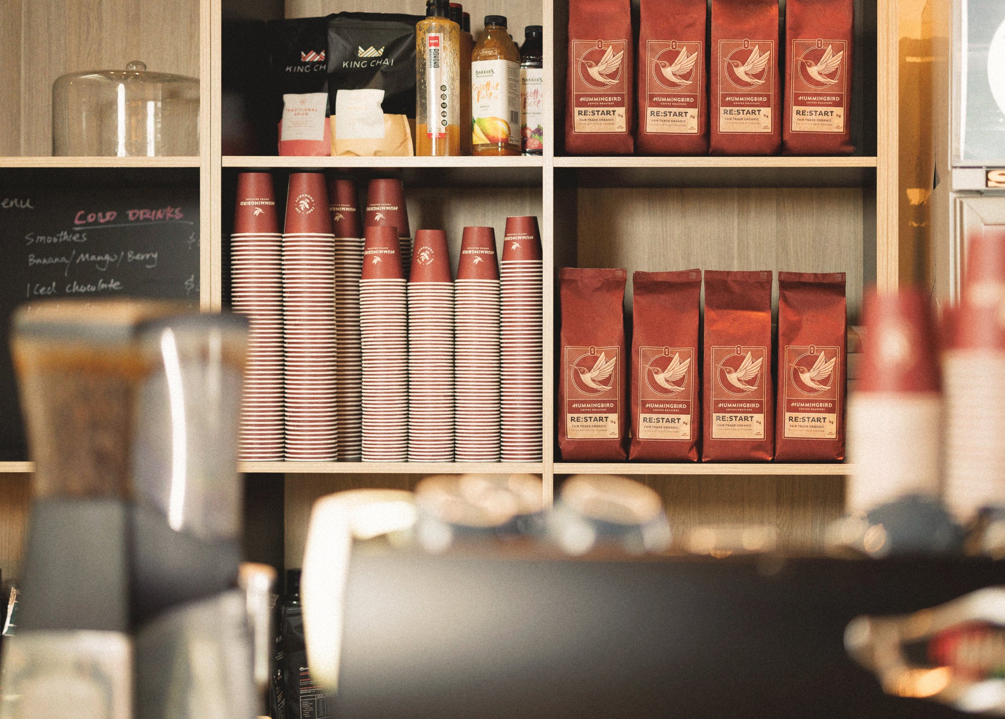 Photo of a cafe stocked with Hummingbird coffee beans and paper cups in front of a coffee machine.