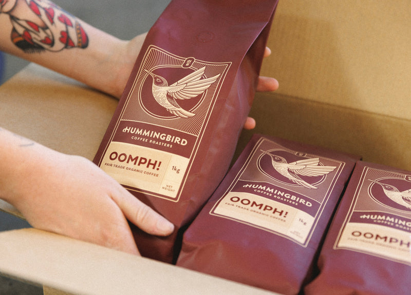 A photo of a pair of hands lifting a package of Hummingbird 'Oomph' Coffee out of a box
